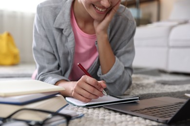 Photo of Smiling student with laptop, notebook and pen preparing for exam on floor indoors, closeup