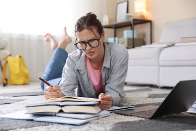 Photo of Worried student in glasses with laptop, notebook and pen preparing for exam on floor indoors