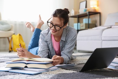 Photo of Smiling student in glasses with laptop, notebook and pen preparing for exam on floor indoors