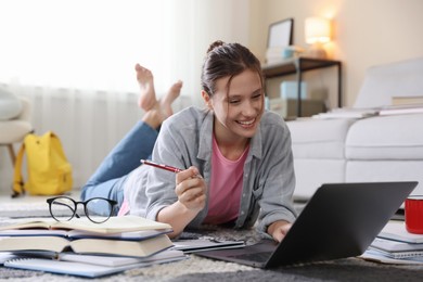 Photo of Smiling student with laptop, books and pen preparing for exam on floor indoors