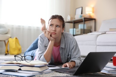 Photo of Worried student with laptop, books and pen preparing for exam on floor indoors