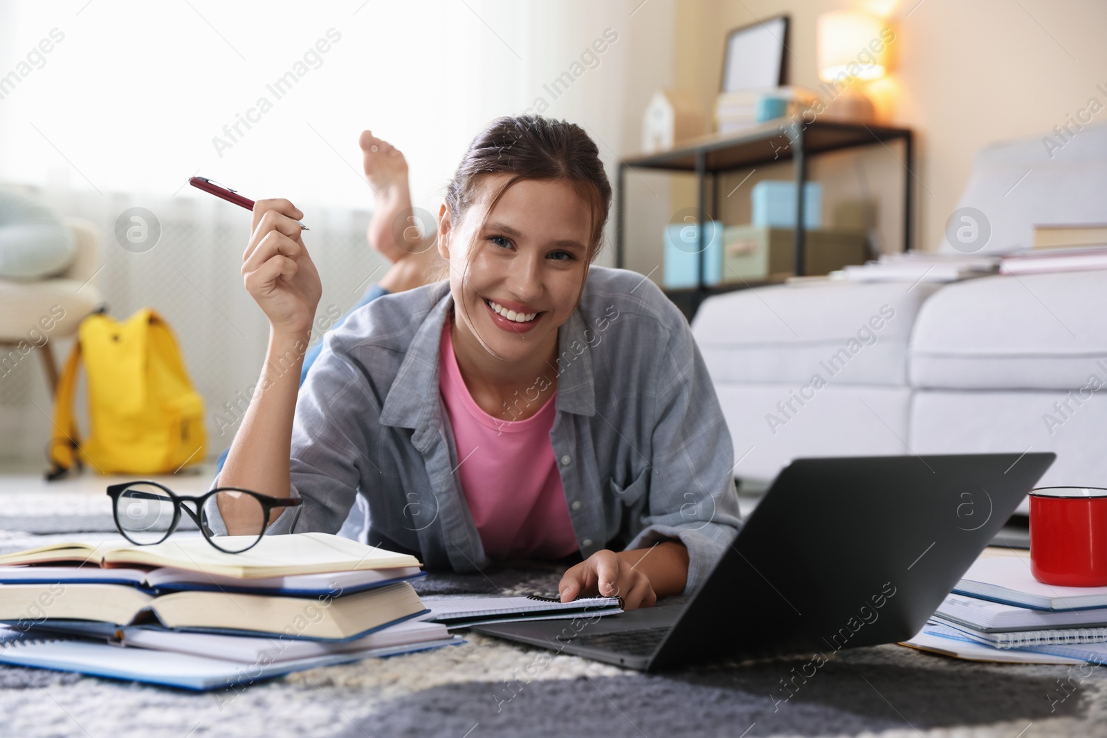 Photo of Smiling student with laptop, books and pen preparing for exam on floor indoors