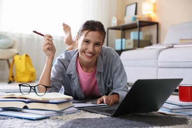 Photo of Smiling student with laptop, books and pen preparing for exam on floor indoors