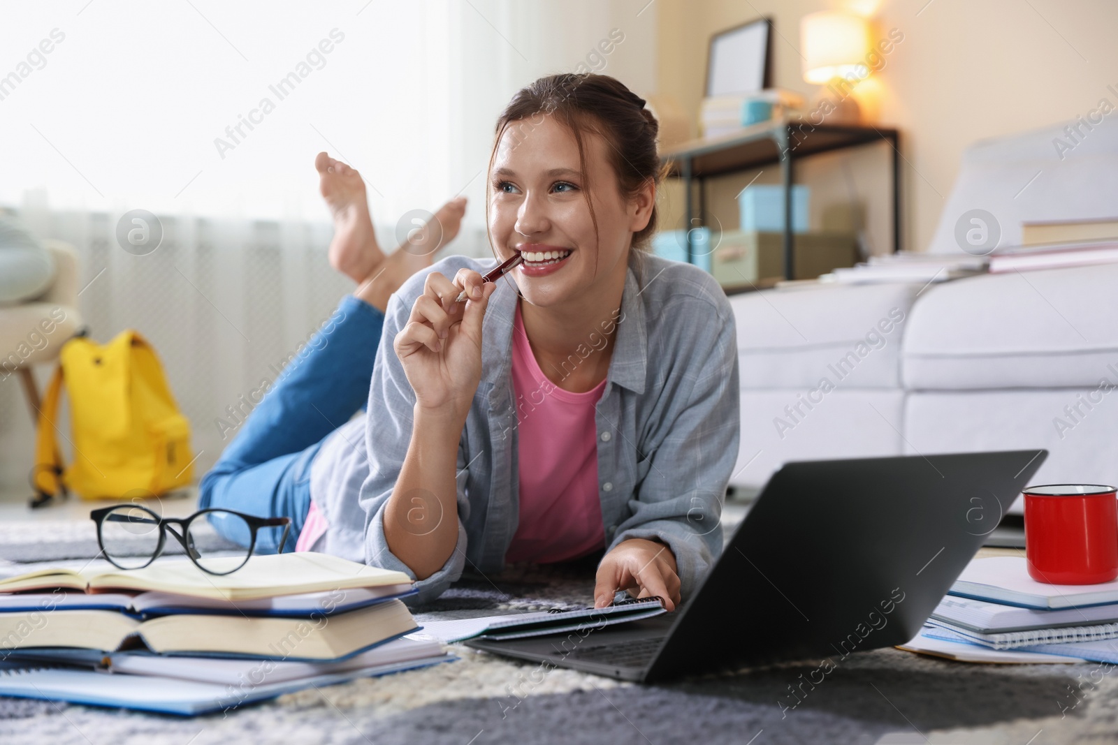 Photo of Smiling student with laptop, books and pen preparing for exam on floor indoors