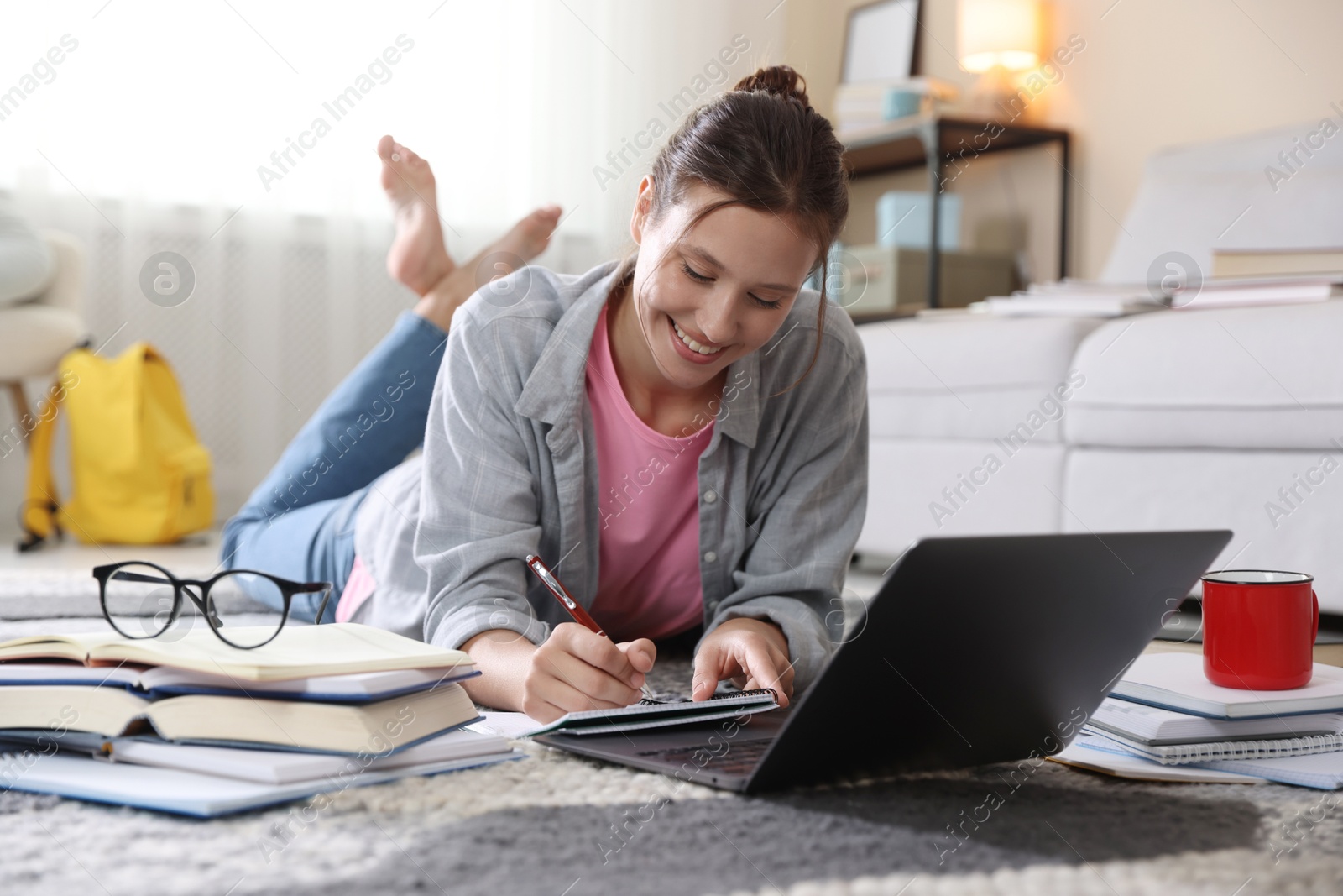 Photo of Smiling student with laptop, books and pen preparing for exam on floor indoors