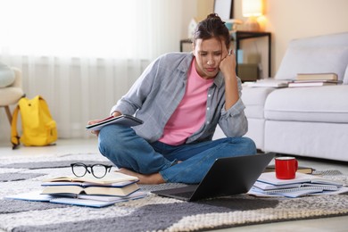 Photo of Worried student with laptop, books and pen preparing for exam on floor indoors