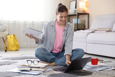 Photo of Smiling student with laptop, books and pen preparing for exam on floor indoors