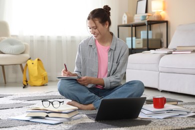 Photo of Smiling student with laptop, books and pen preparing for exam on floor indoors