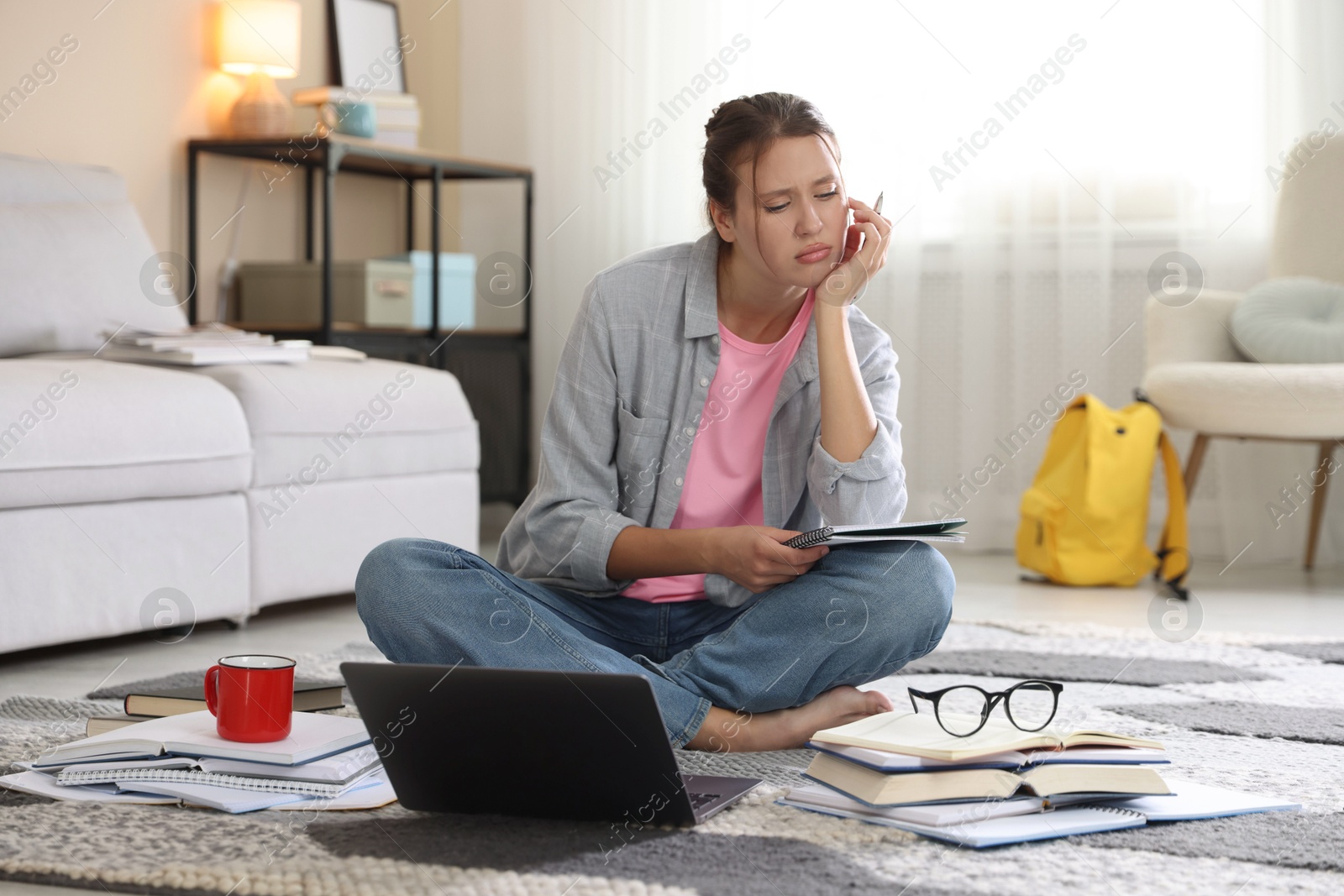 Photo of Worried student with laptop, books and pen preparing for exam on floor indoors
