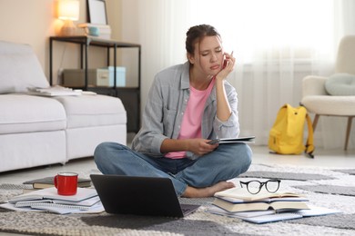 Photo of Worried student with laptop, books and pen preparing for exam on floor indoors
