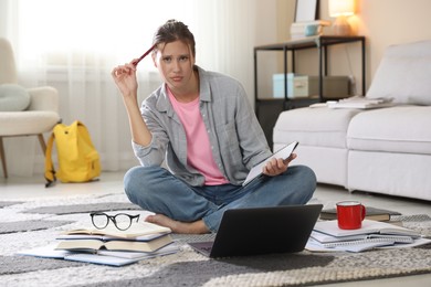 Photo of Worried student with laptop, books and pen preparing for exam on floor indoors