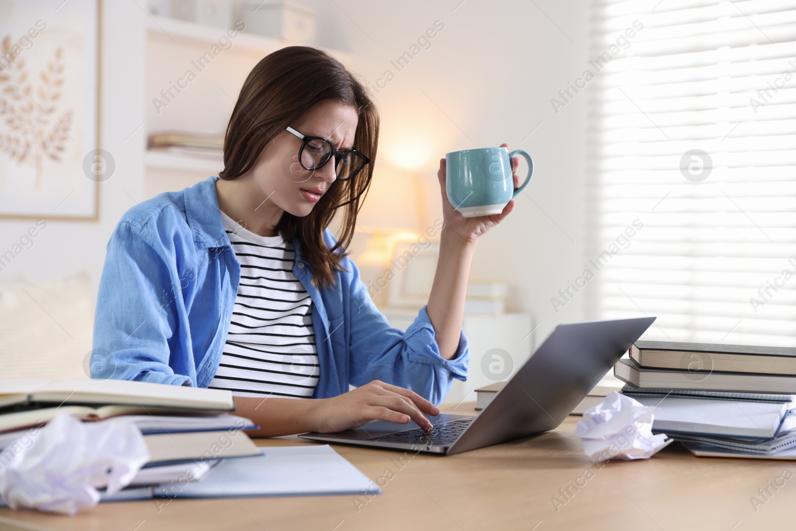 Photo of Worried student in glasses with laptop, books and cup preparing for exam at table indoors