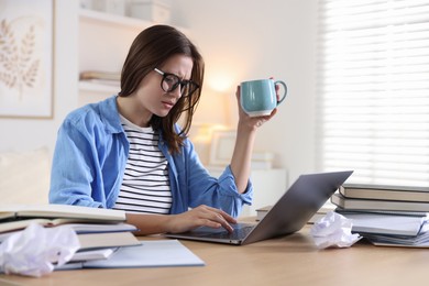 Photo of Worried student in glasses with laptop, books and cup preparing for exam at table indoors