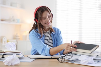 Photo of Smiling student in headphones with pen and books preparing for exam at table indoors