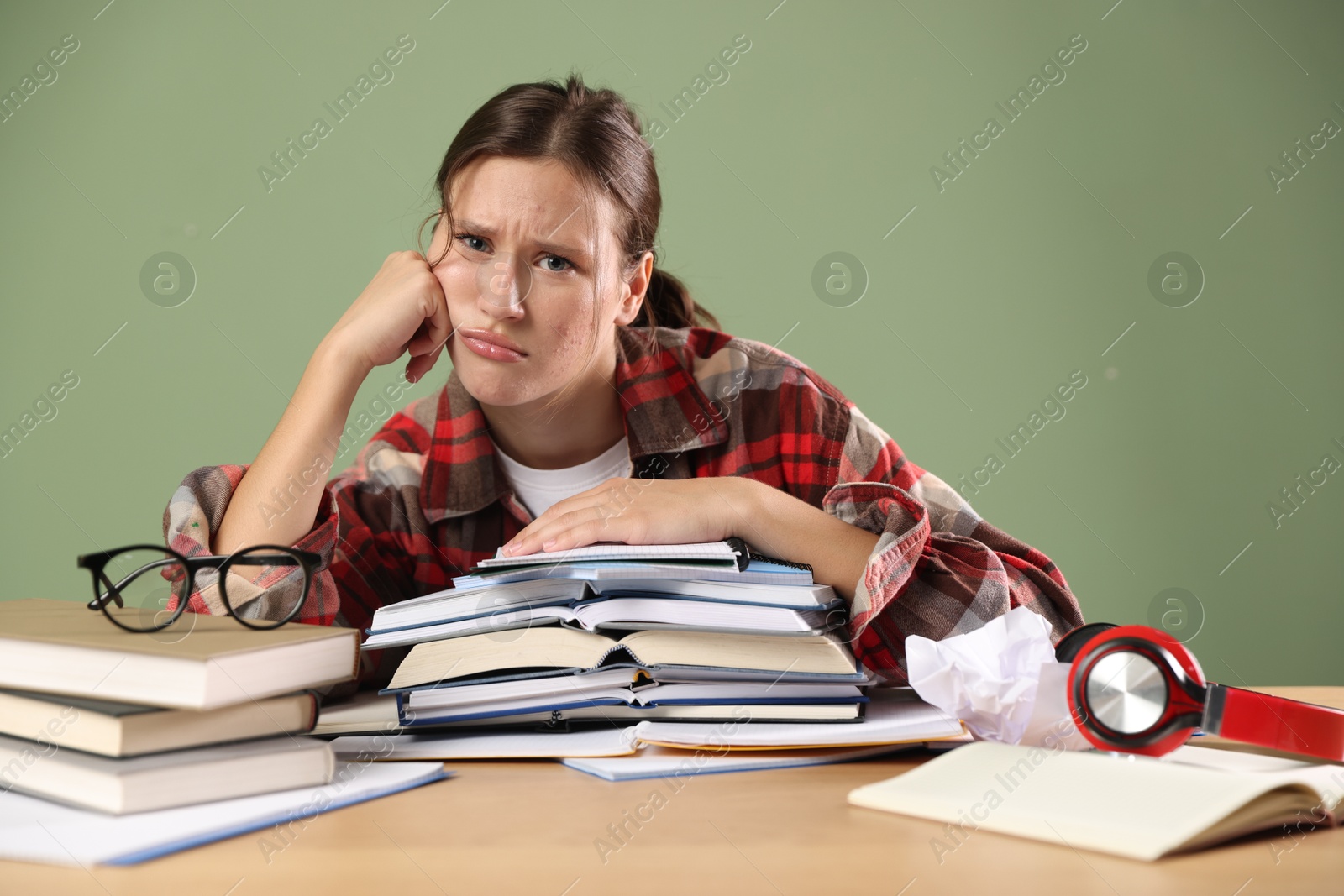 Photo of Tired student with books preparing for exam at wooden table near green wall