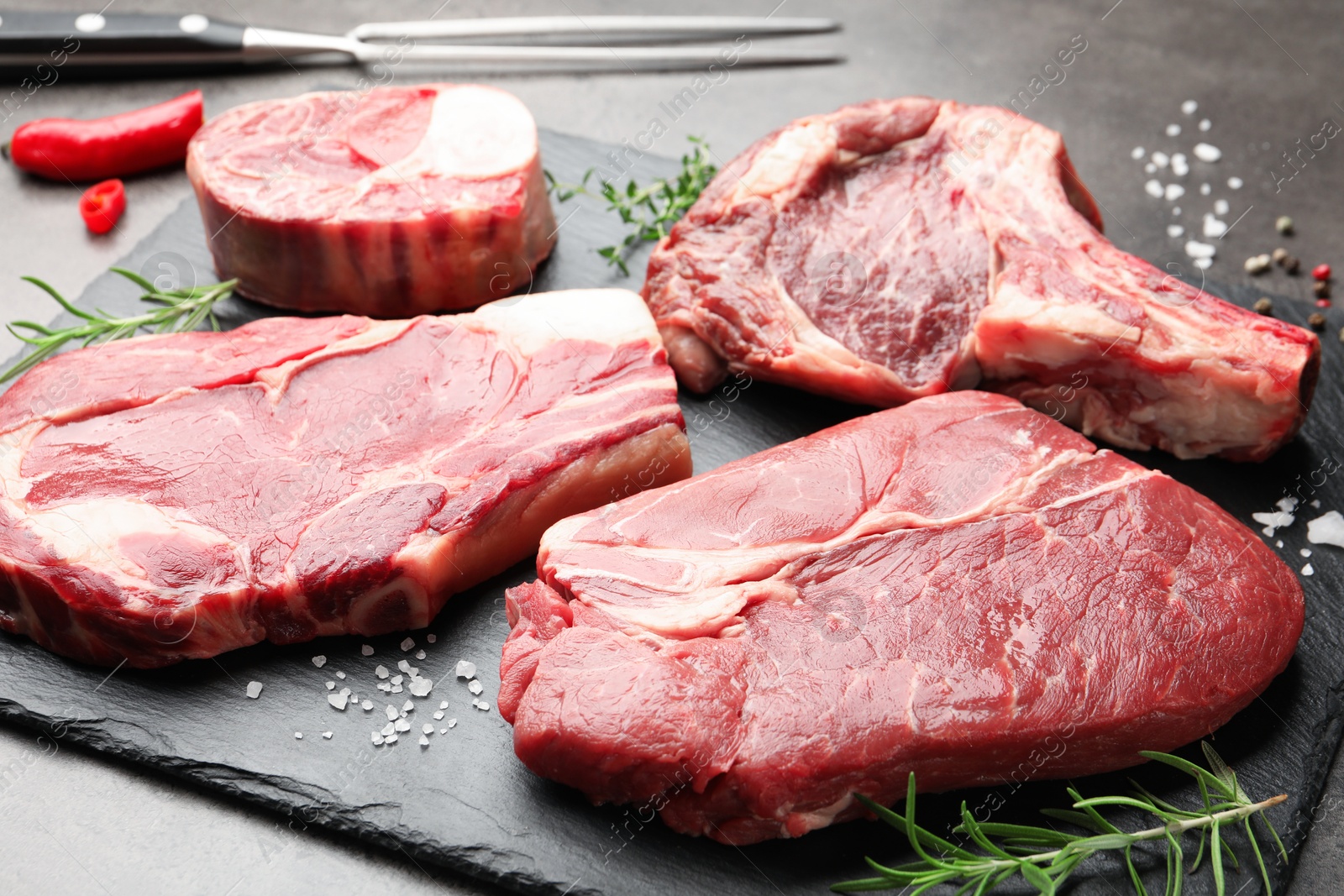 Pieces of raw beef meat, spices and carving fork on grey table, closeup Photo of Pieces of raw beef meat, spices and carving fork on grey table, closeup