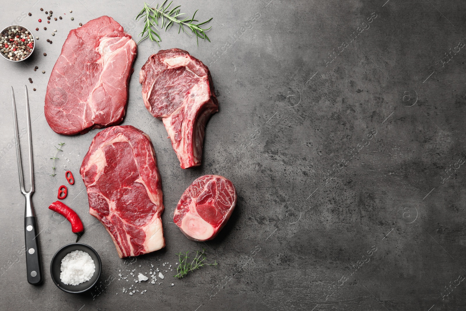 Pieces of raw beef meat, spices and carving fork on grey table, flat lay. Space for text Photo of Pieces of raw beef meat, spices and carving fork on grey table, flat lay. Space for text