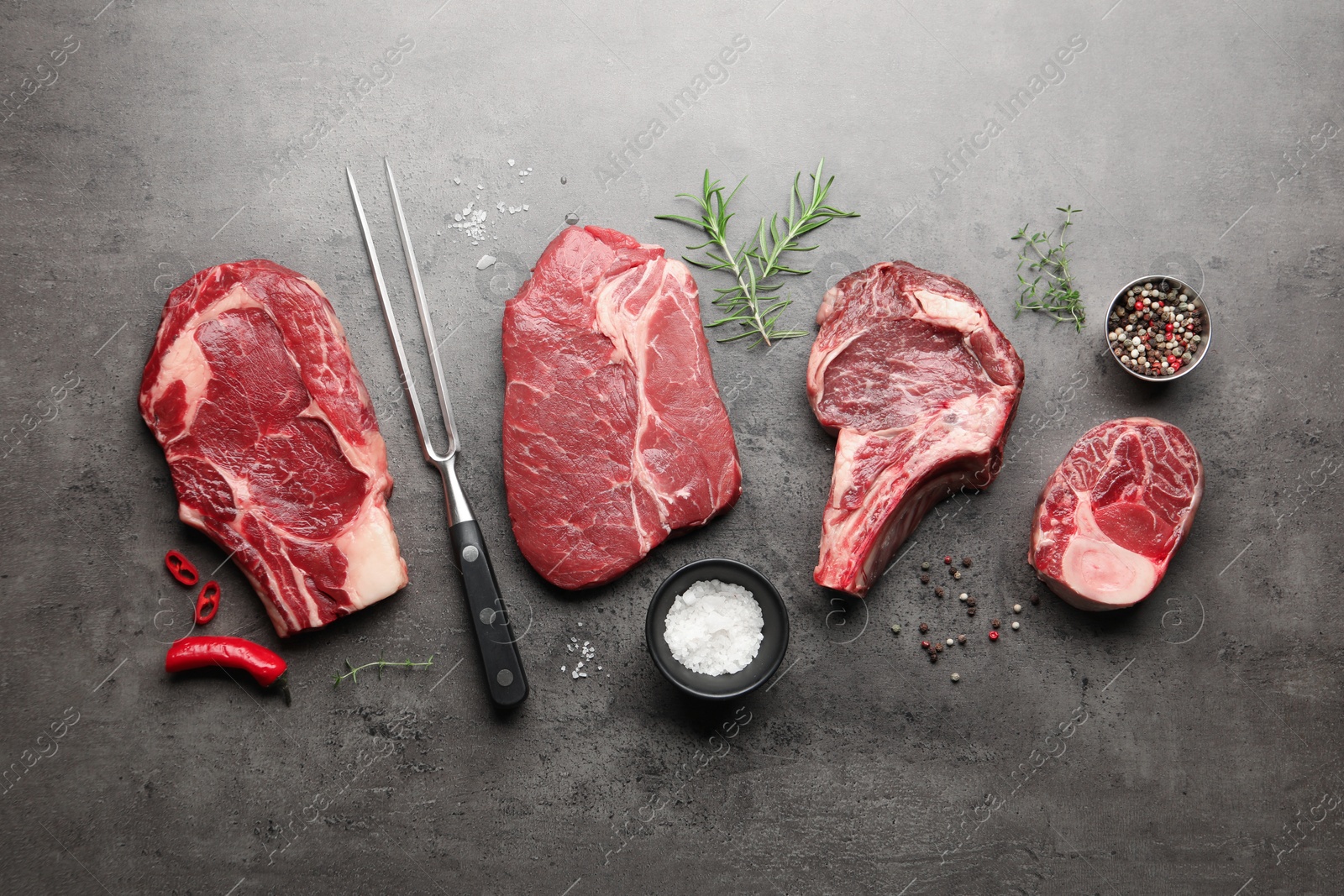 Pieces of raw beef meat, spices and carving fork on grey table, flat lay Photo of Pieces of raw beef meat, spices and carving fork on grey table, flat lay
