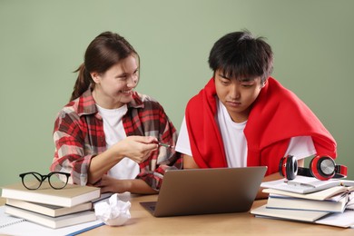 Photo of Happy students with laptop and books preparing for exam at wooden table near green wall