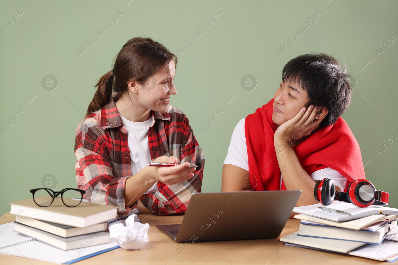 Photo of Happy students with laptop and books preparing for exam at wooden table near green wall