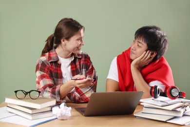 Photo of Happy students with laptop and books preparing for exam at wooden table near green wall