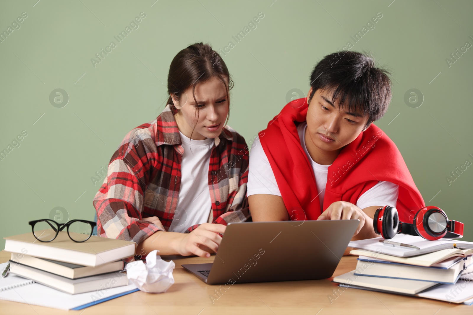 Photo of Students with laptop and books preparing for exam at wooden table near green wall