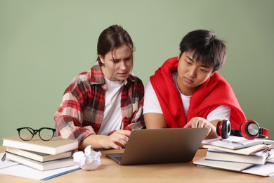 Photo of Students with laptop and books preparing for exam at wooden table near green wall
