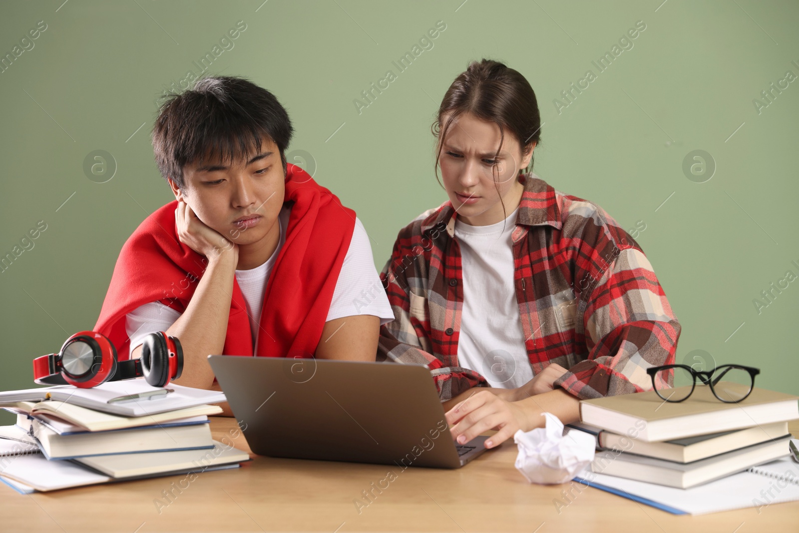 Photo of Students with laptop and books preparing for exam at wooden table near green wall