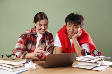 Photo of Happy students with laptop and books preparing for exam at wooden table near green wall