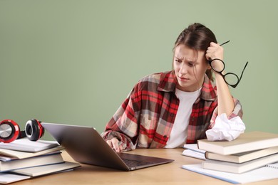 Photo of Worried student with glasses, laptop and books preparing for exam at wooden table near green wall