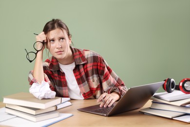 Photo of Worried student with glasses, laptop and books preparing for exam at wooden table near green wall