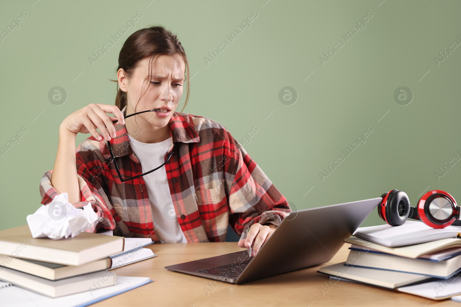 Photo of Worried student with glasses, laptop and books preparing for exam at wooden table near green wall