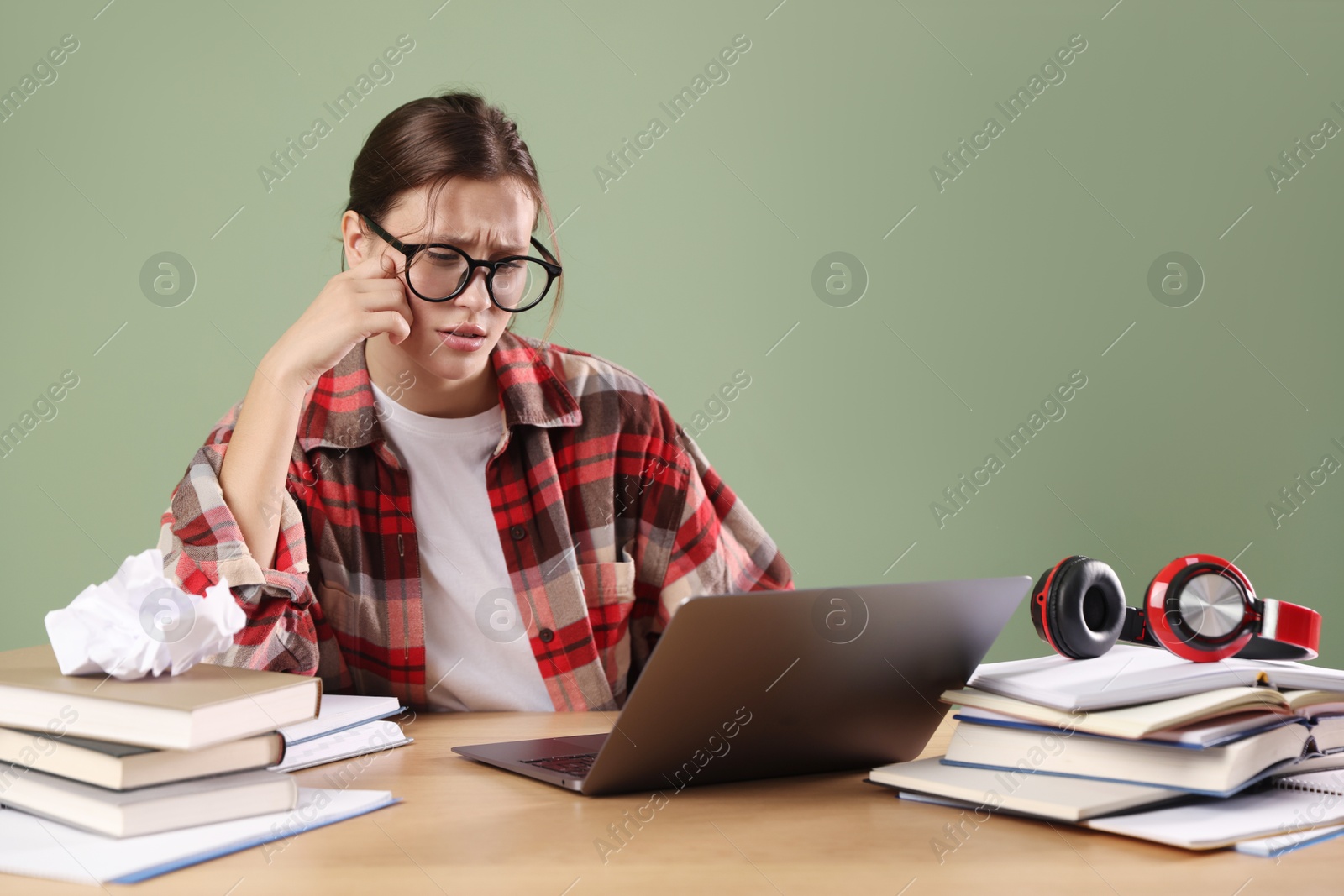 Photo of Worried student in glasses with laptop and books preparing for exam at wooden table near green wall