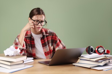 Photo of Worried student in glasses with laptop and books preparing for exam at wooden table near green wall