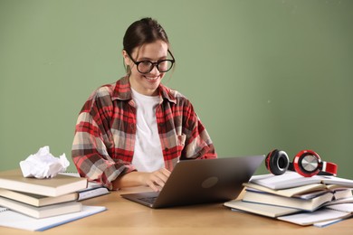 Photo of Smiling student in glasses with laptop and books preparing for exam at wooden table near green wall