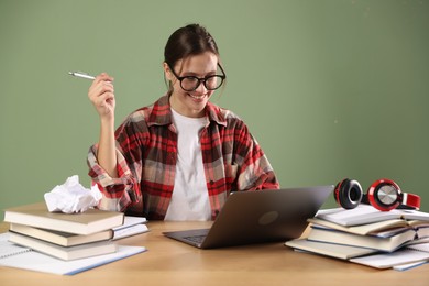 Photo of Smiling student in glasses with laptop, pen and books preparing for exam at wooden table near green wall