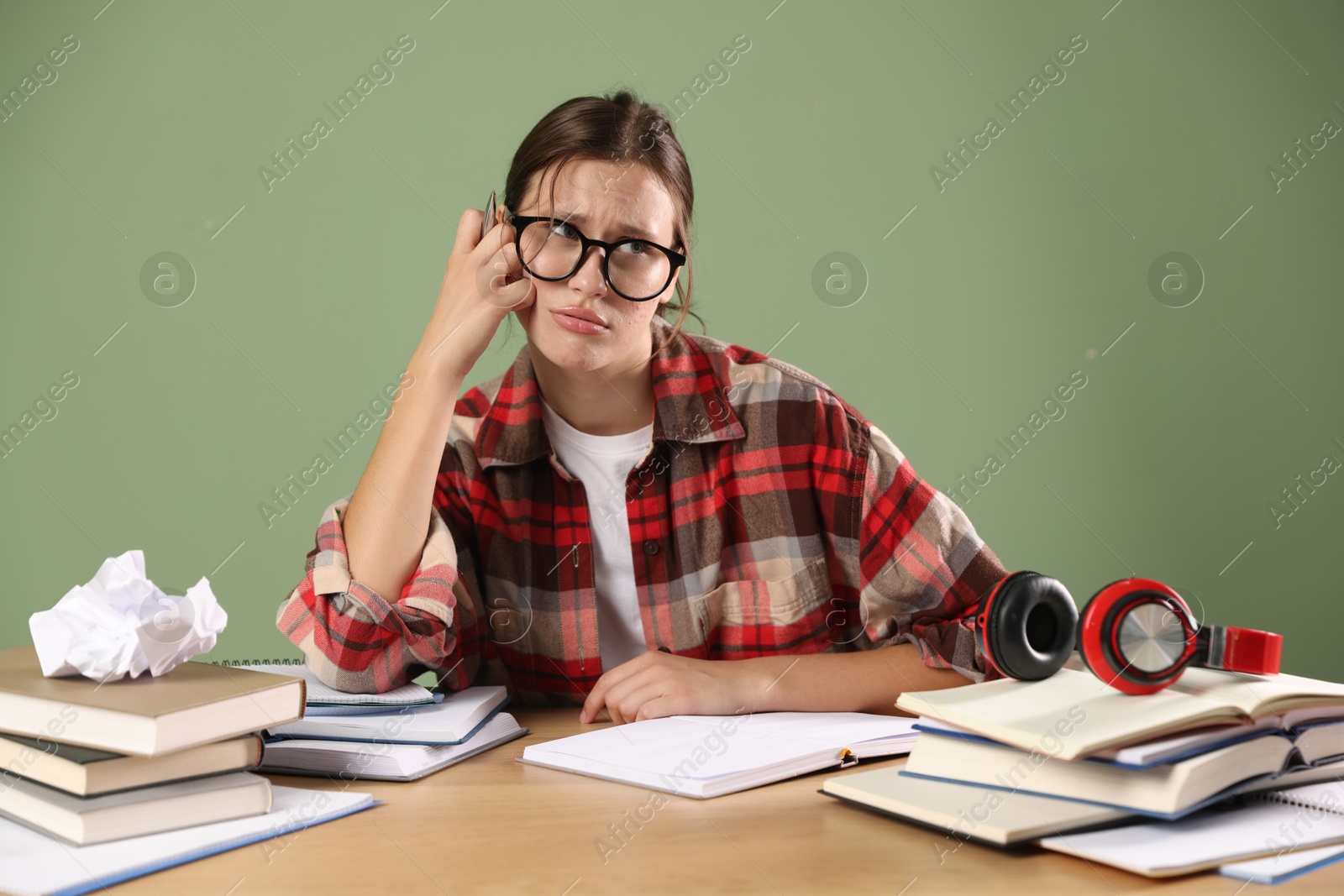 Photo of Worried student in glasses with pen and books preparing for exam at wooden table near green wall