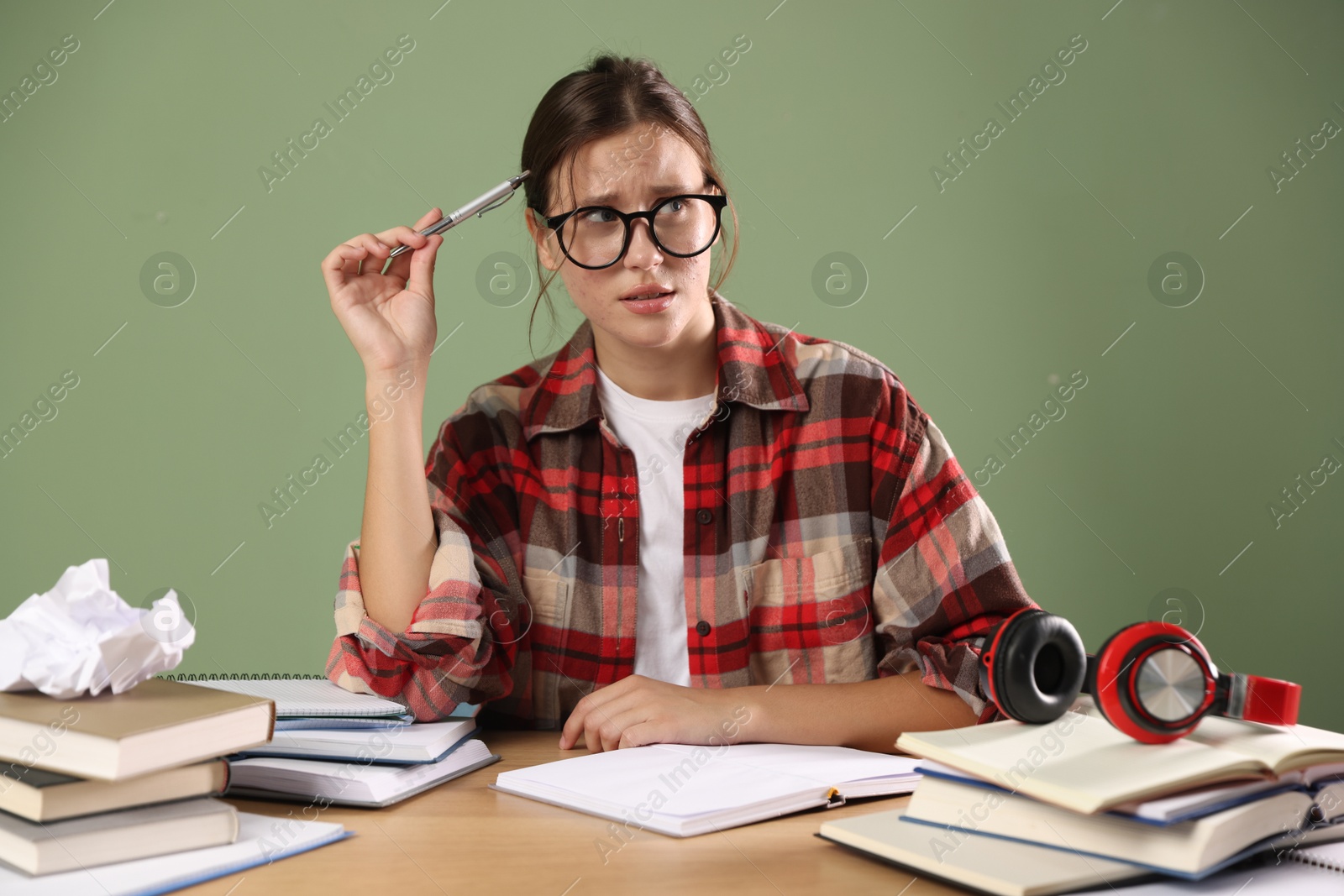 Photo of Worried student in glasses with pen and books preparing for exam at wooden table near green wall