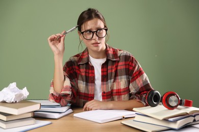 Photo of Worried student in glasses with pen and books preparing for exam at wooden table near green wall