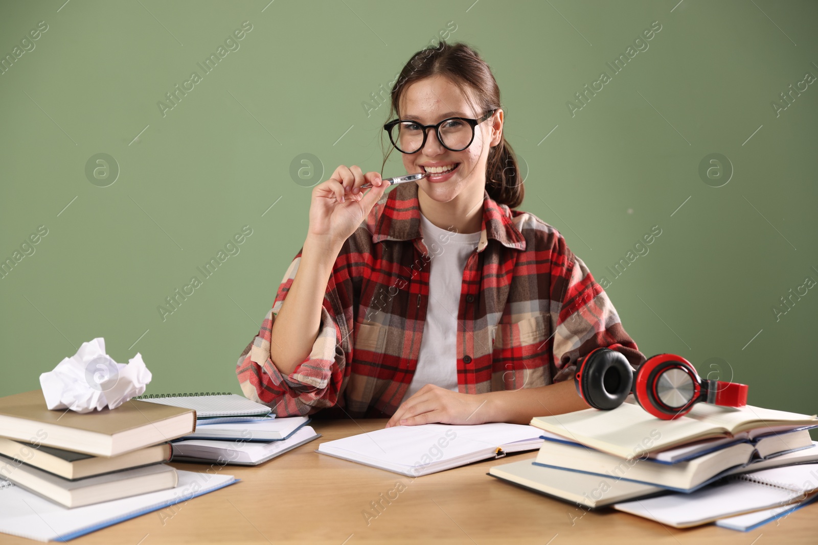 Photo of Smiling student in glasses with pen and books preparing for exam at wooden table near green wall