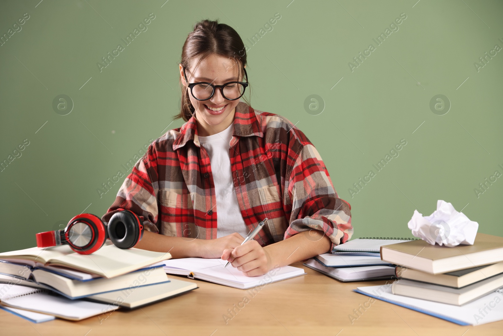 Photo of Smiling student in glasses with pen and books preparing for exam at wooden table near green wall