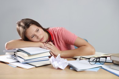 Photo of Tired student with books preparing for exam at wooden table near grey wall
