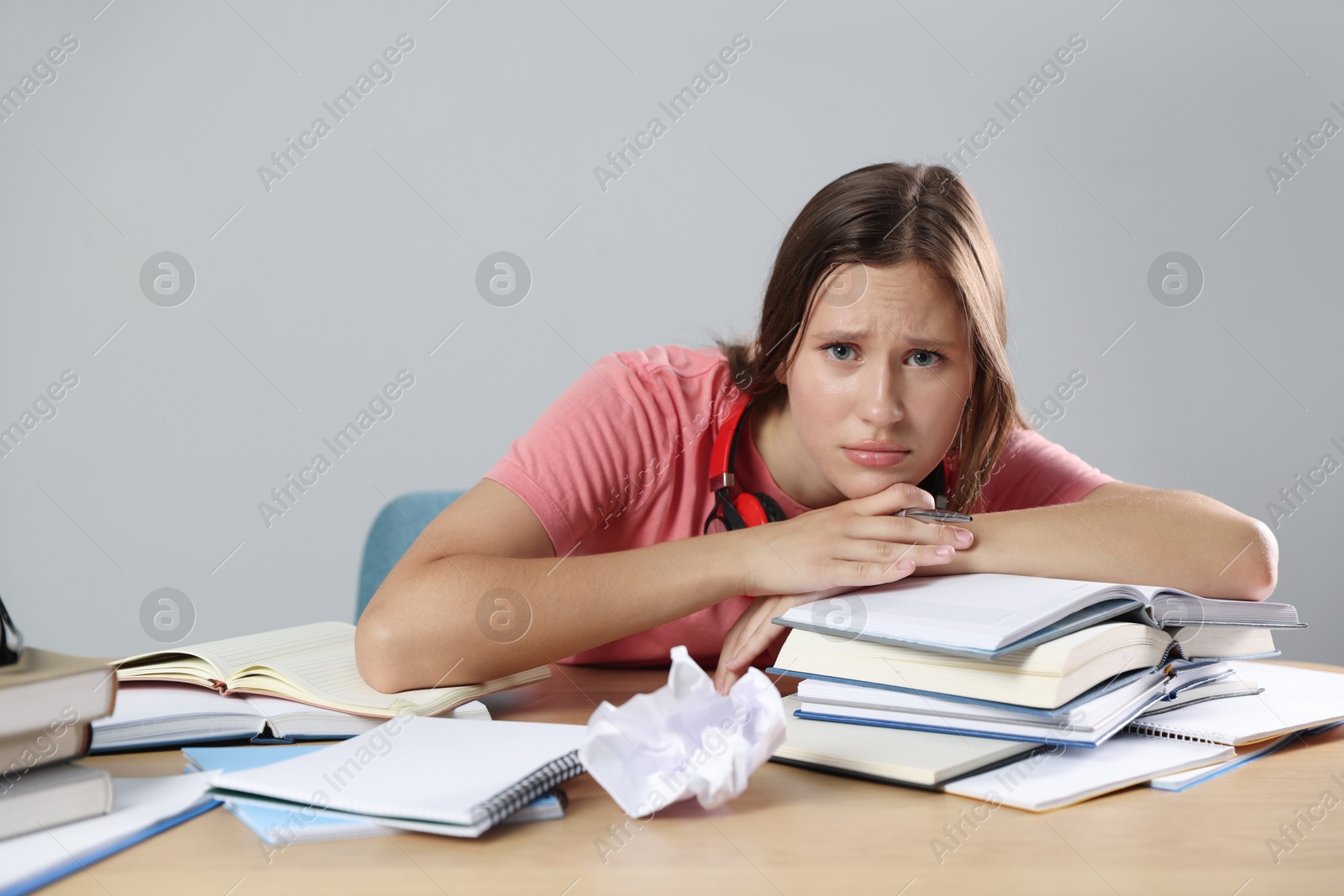 Photo of Tired student with books preparing for exam at wooden table near grey wall