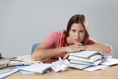 Photo of Tired student with books preparing for exam at wooden table near grey wall