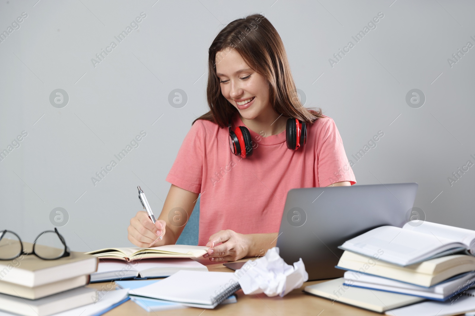 Photo of Smiling student with laptop, pen and books preparing for exam at table near grey wall