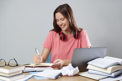 Photo of Smiling student with laptop, pen and books preparing for exam at table near grey wall