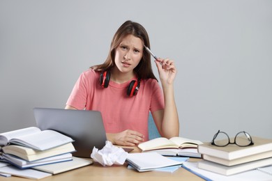 Photo of Tired student with laptop, pen and books preparing for exam at table near grey wall