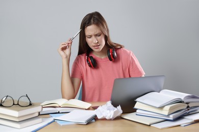 Photo of Tired student with laptop, pen and books preparing for exam at table near grey wall