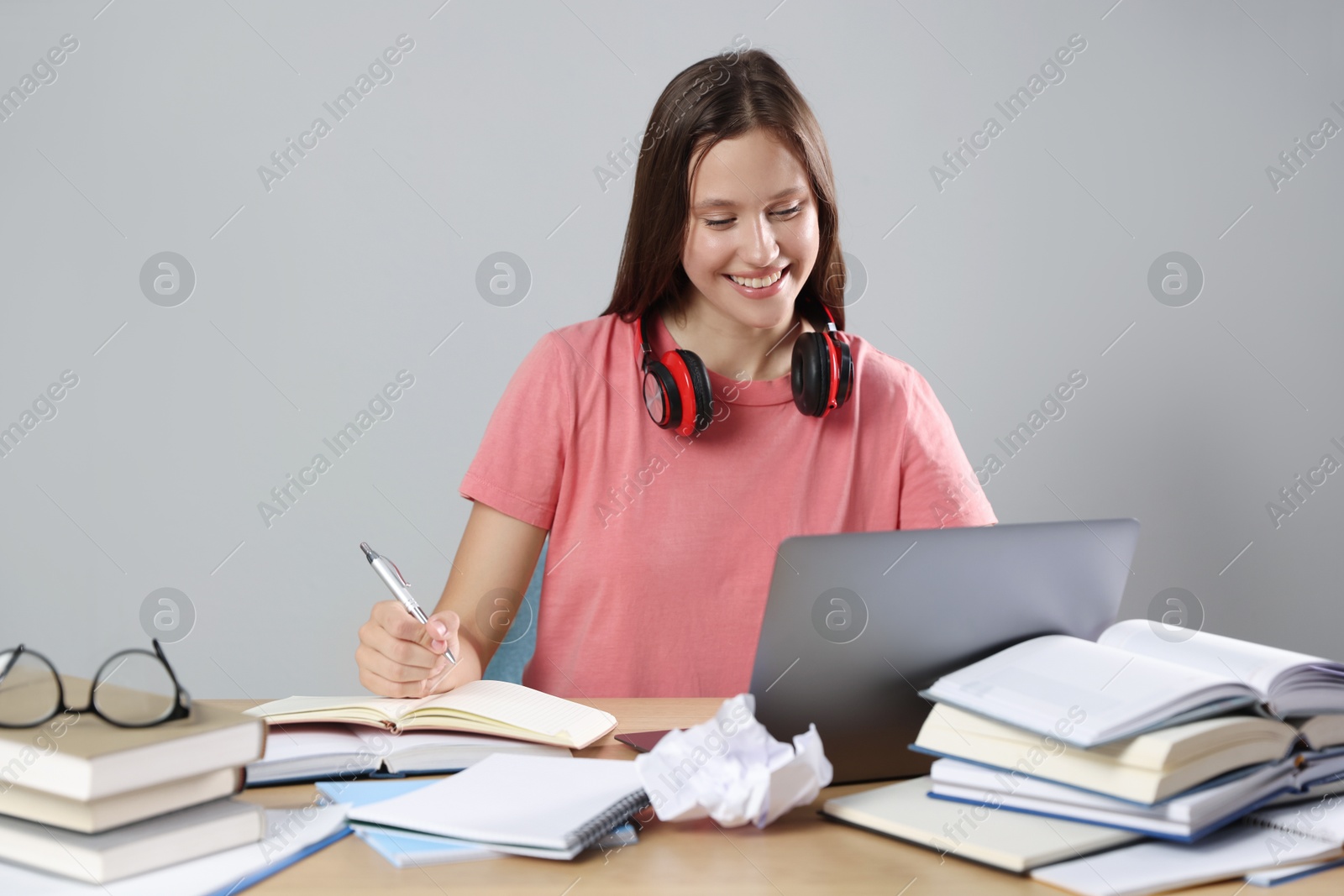 Photo of Smiling student with laptop, pen and books preparing for exam at table near grey wall