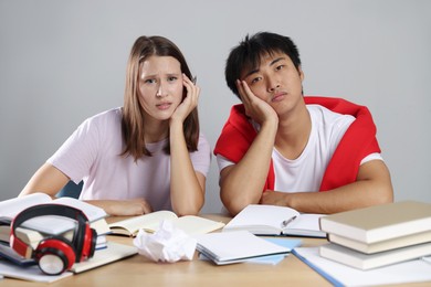 Photo of Tired students with books preparing for exam at table near grey wall